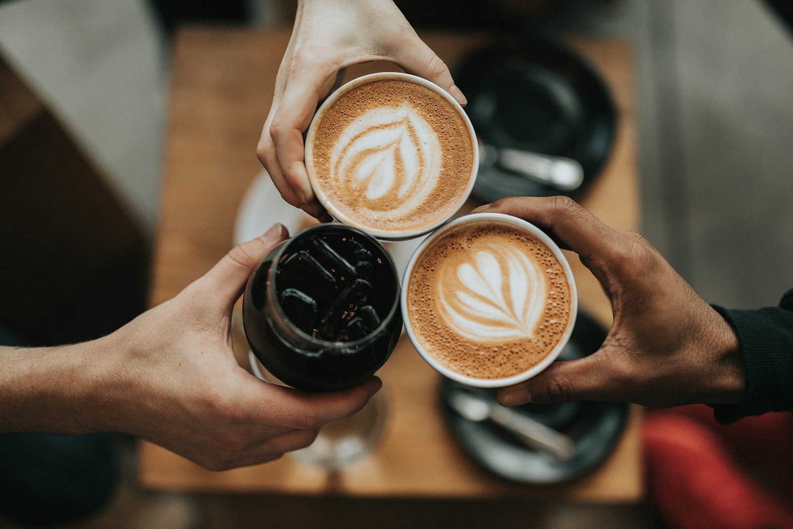 Three friends raising latte cups and an iced coffee together over a cafe table
