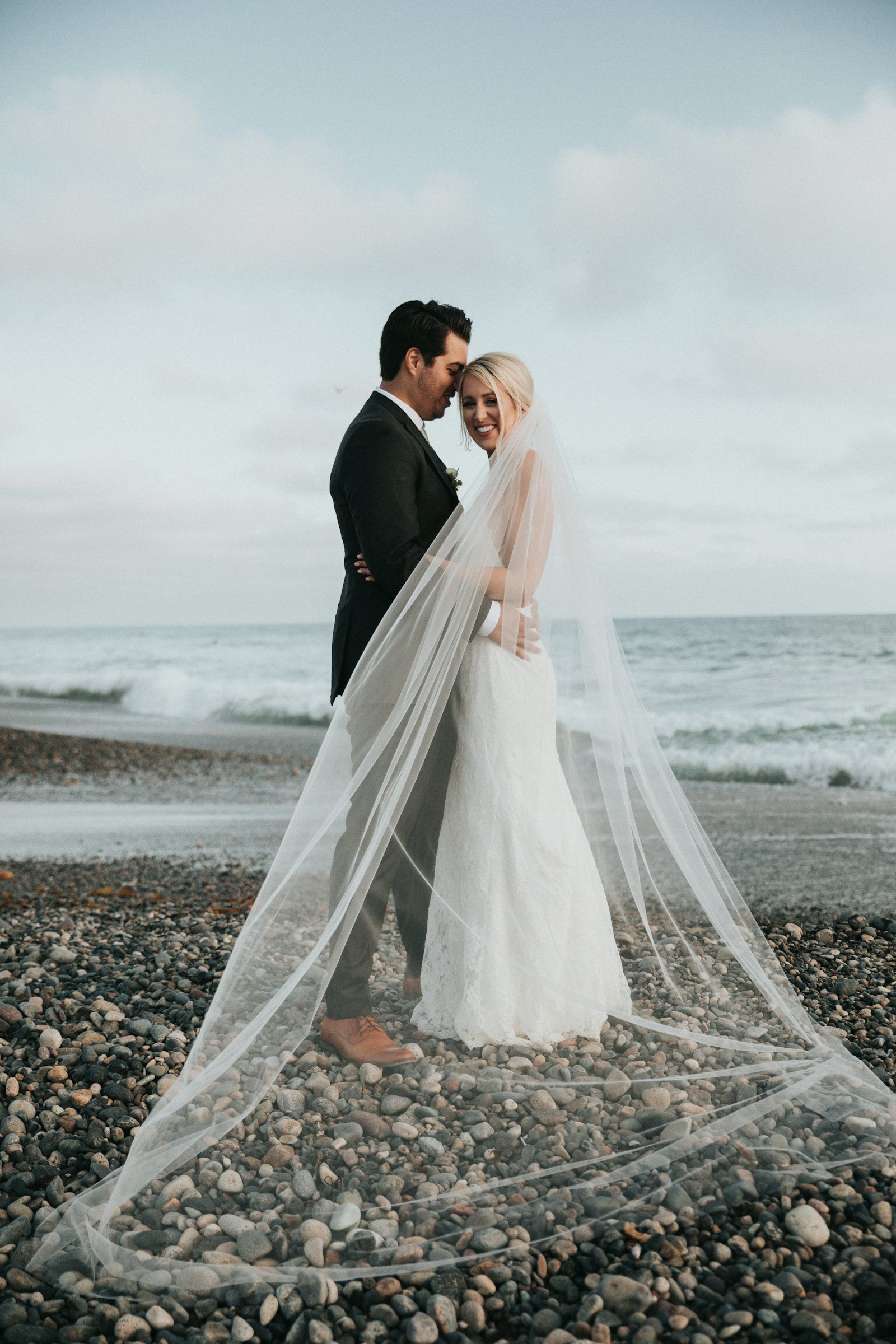 Bride and groom embracing on a pebbled beach, veil trailing in the sea breeze