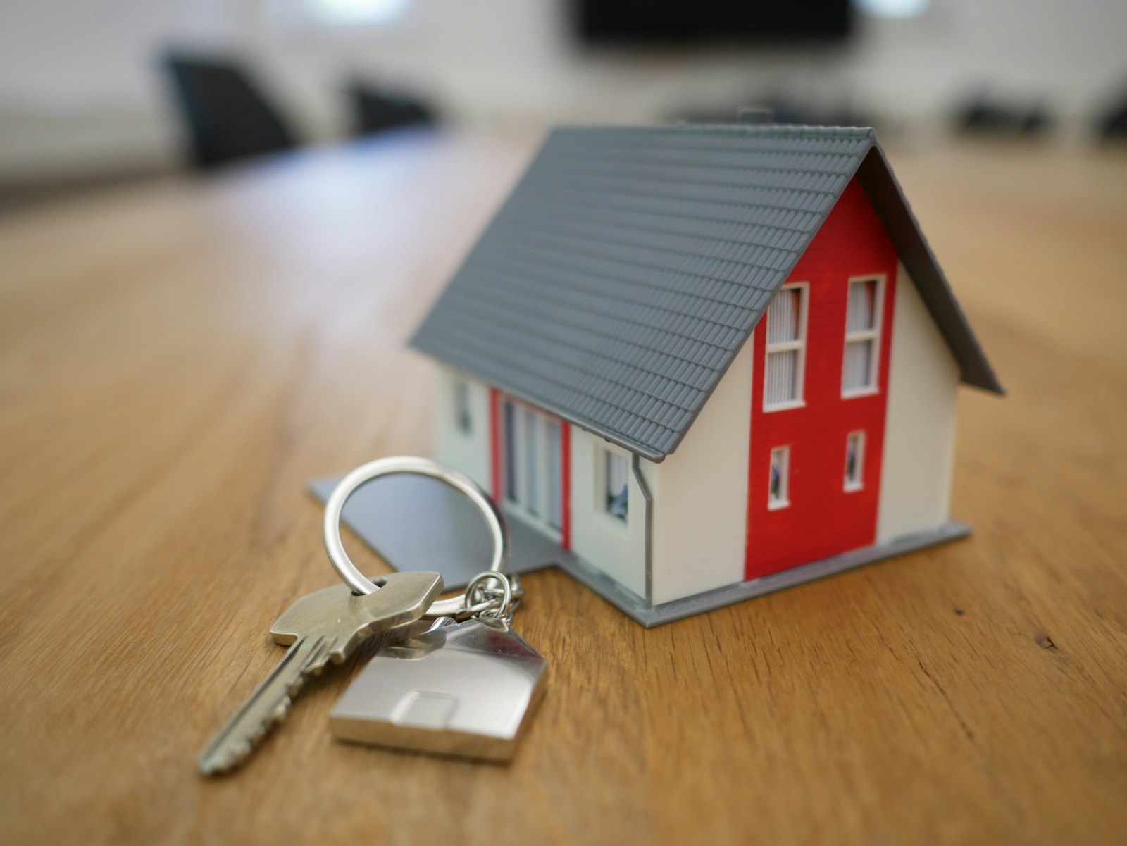 House keys with a home-shaped keyring beside a miniature house on a wooden table