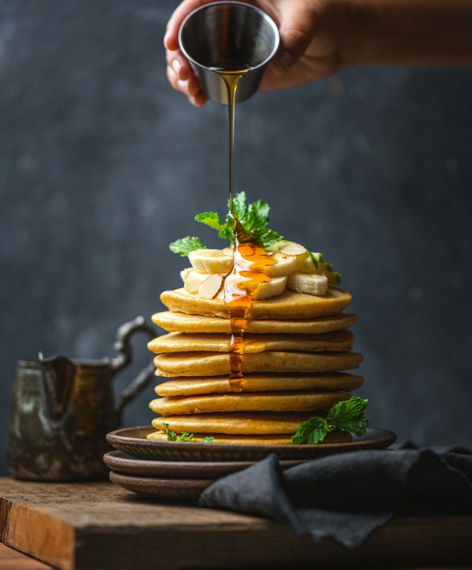 Golden syrup being poured over a tall stack of pancakes with banana and mint