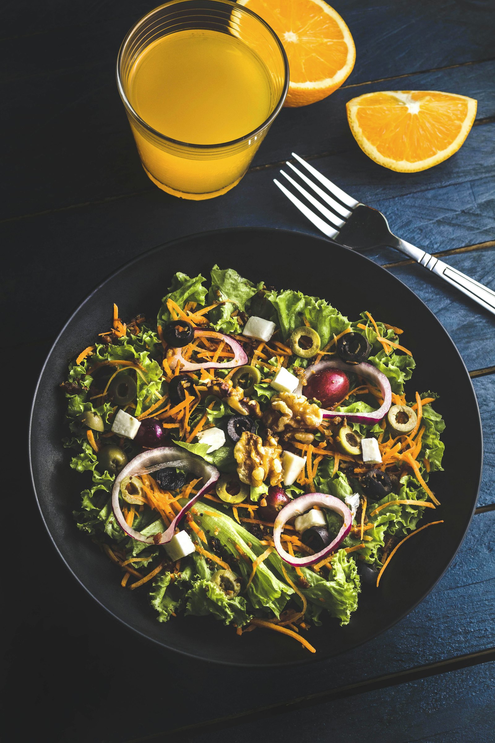 Dark plate of garden salad with olives, feta and red onion beside fresh orange juice