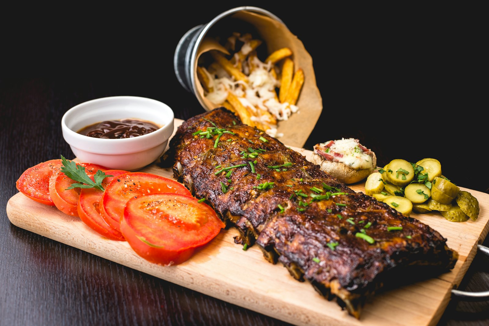 Charred barbecue ribs on a wooden board with tomatoes, pickles and smoky sauce
