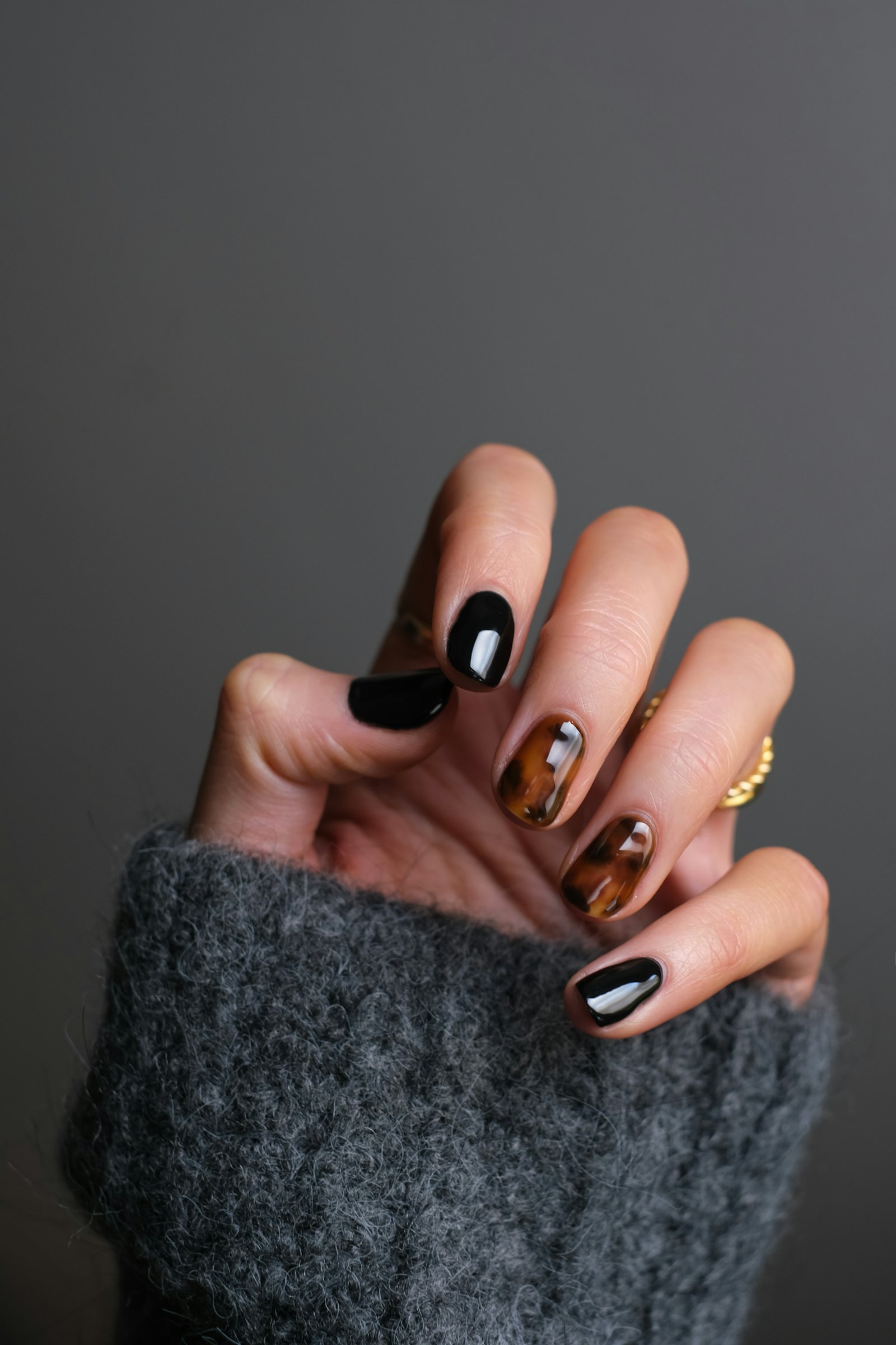 Hand with glossy black and tortoiseshell gel nail art against a grey knit sleeve
